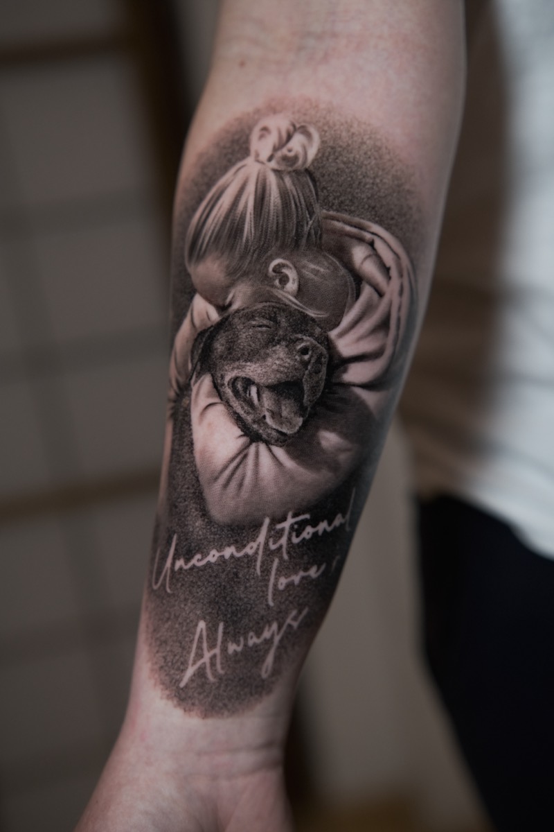 Portrait-Tattoo am Unterarm in Black & Grey: ein Kind umarmt einen lachenden Hund, darunter der Schriftzug „Unconditional love, Always“