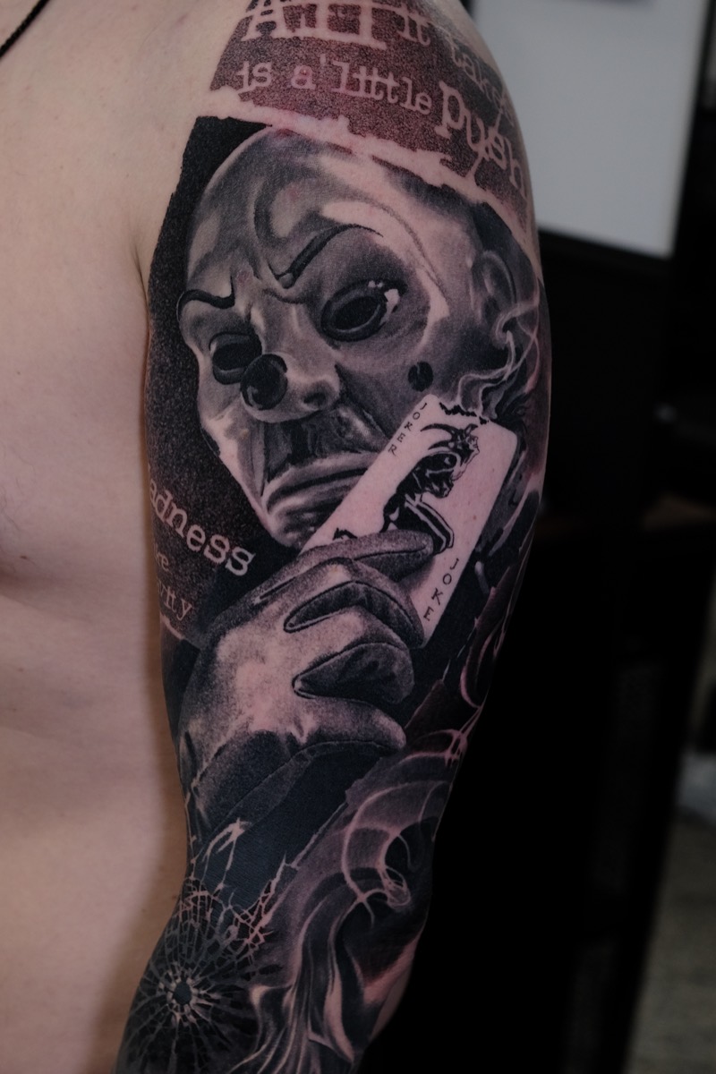 Black & Grey Tattoo am Oberarm: Figur mit Clownsmaske hält eine Joker-Spielkarte, Schrift-Collage und Rauch