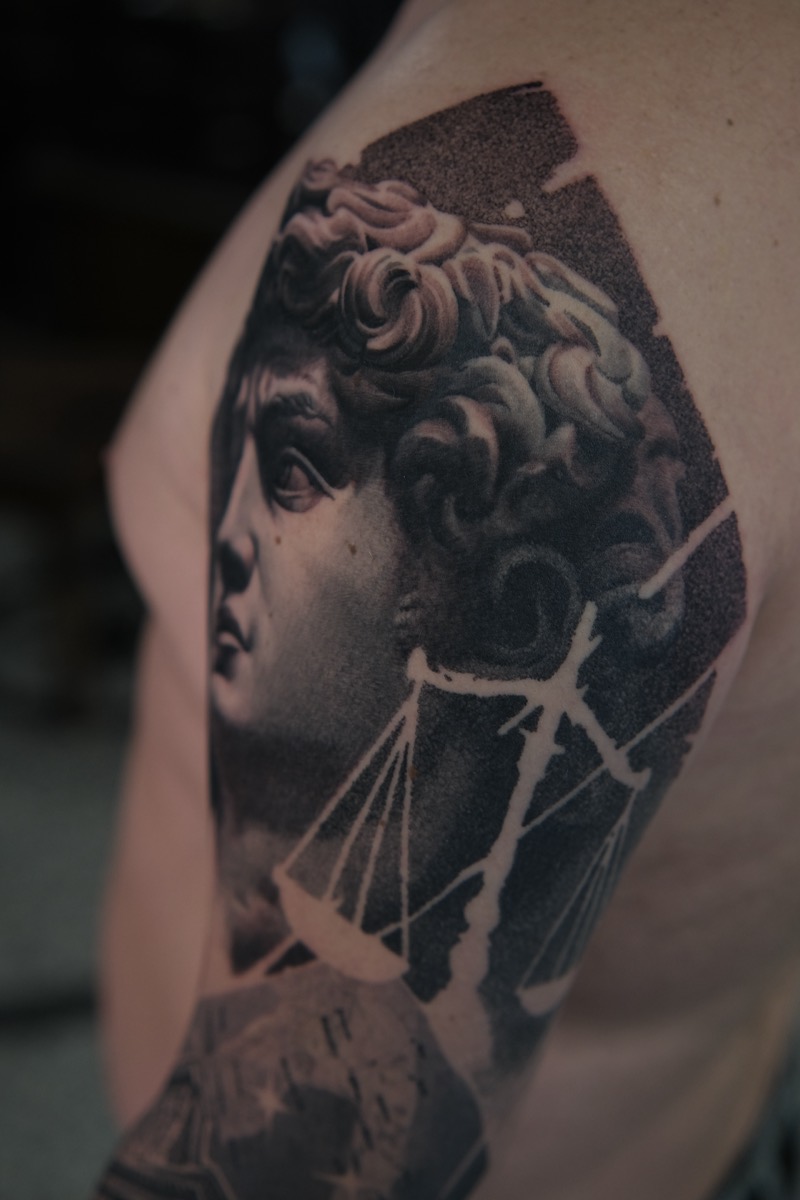 Detailansicht am Oberarm: Kopf der David-Statue im Profil mit Waage, Dotwork-Hintergrund
