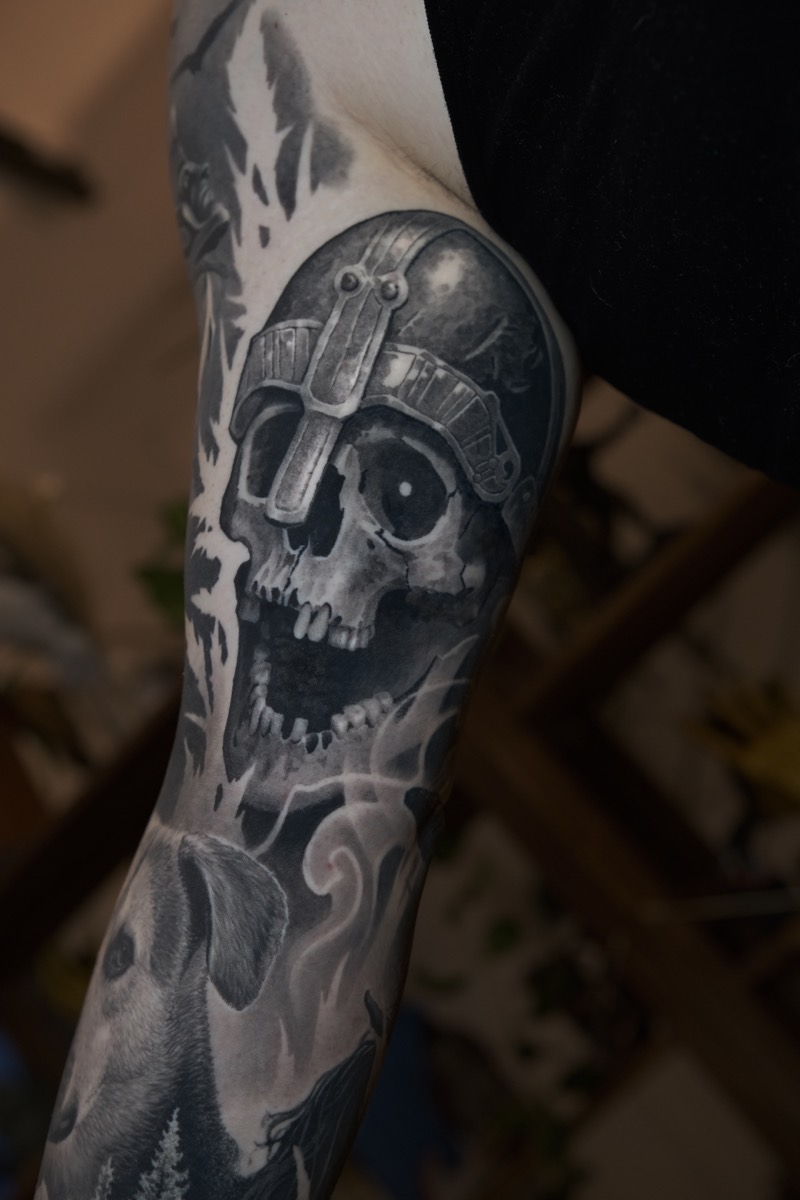 Black & Grey Tattoo am Arm: Totenkopf mit Helm, Rauch und darunter ein Hundekopf