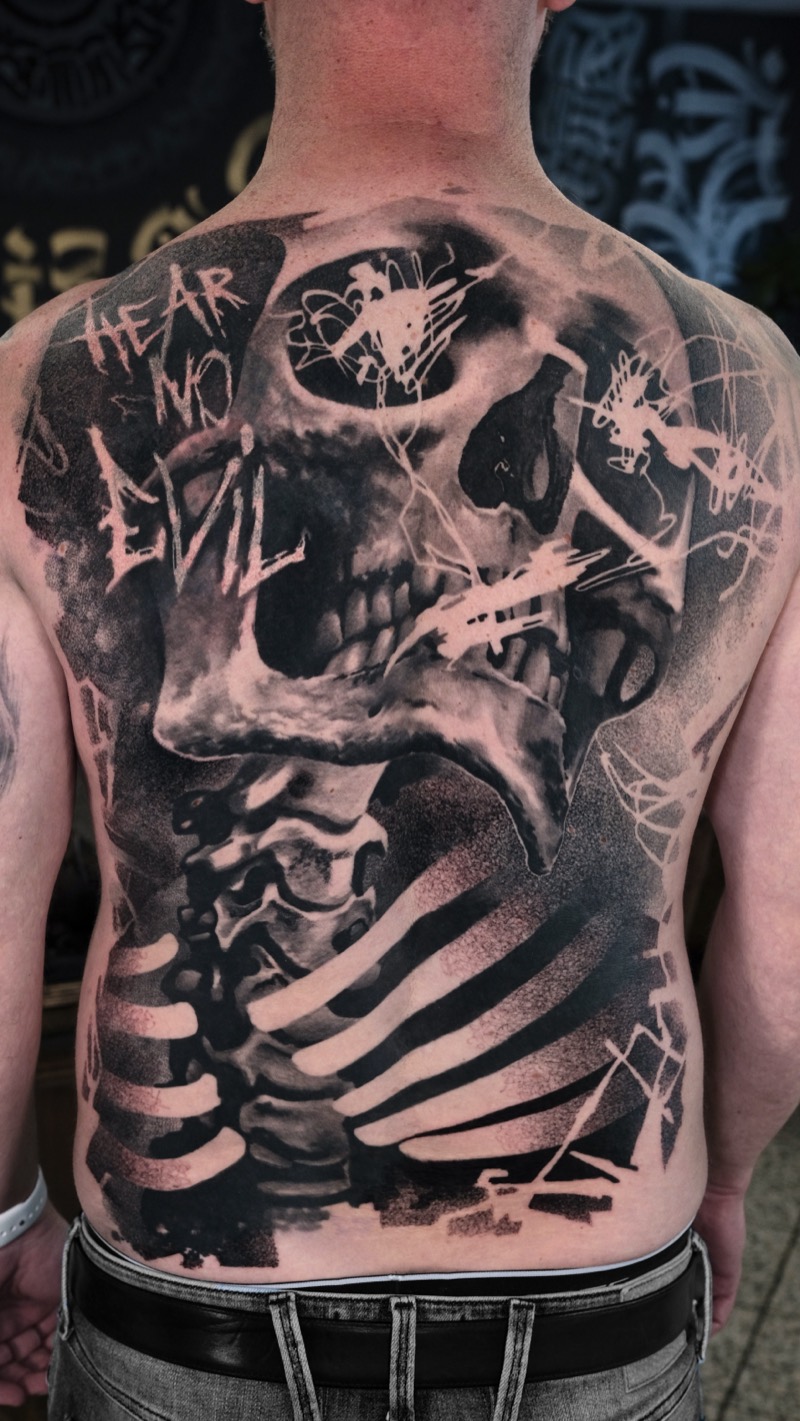 Rücken-Tattoo in Black & Grey: großer Totenkopf mit Wirbelsäule und Rippen, Schriftzug „Hear no evil“ und grafische Pinselstrich-Elemente