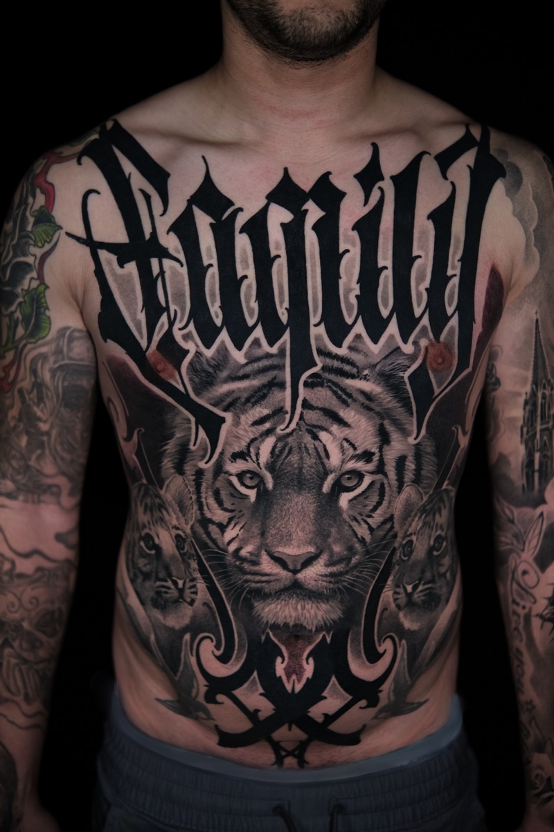 Großflächiges Tattoo auf Brust und Bauch: realistischer Tigerkopf mit Tigerjungen unter einem Blackletter-Schriftzug „Family“