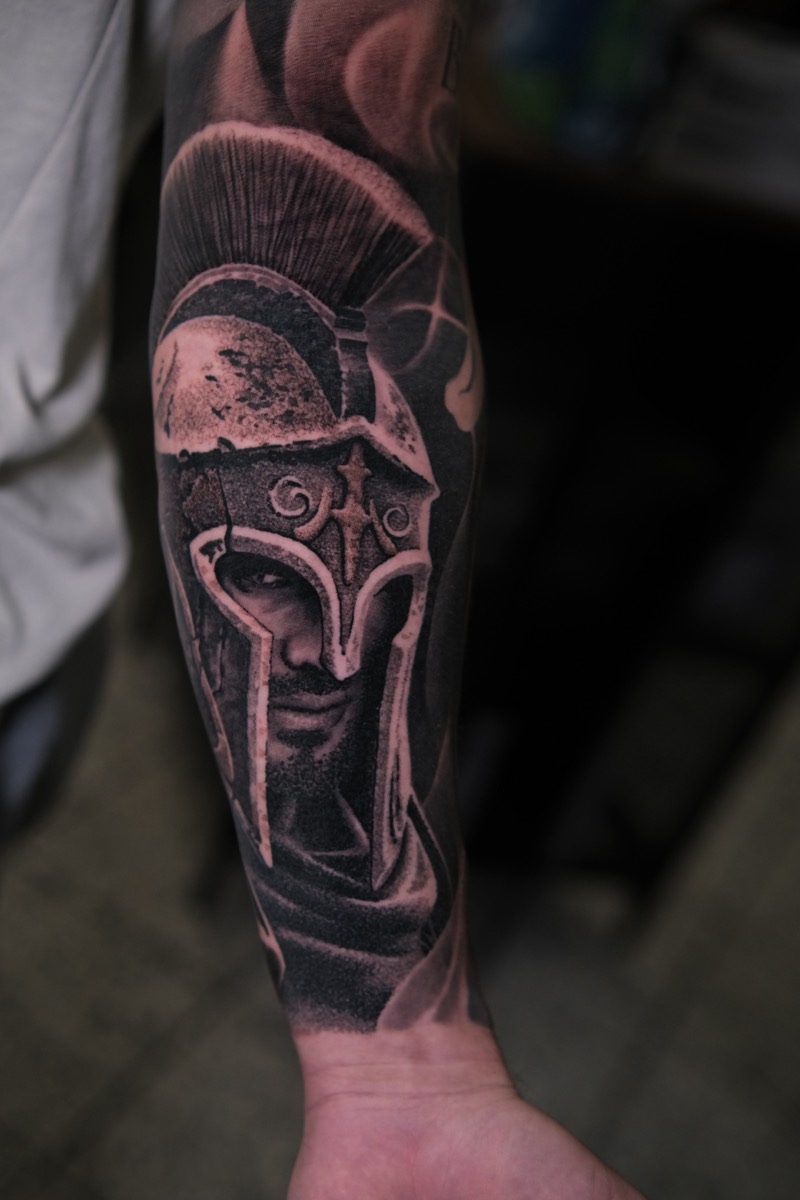Black & Grey Tattoo am Unterarm: Krieger mit korinthischem Helm, Blick aus dem Schatten des Helms